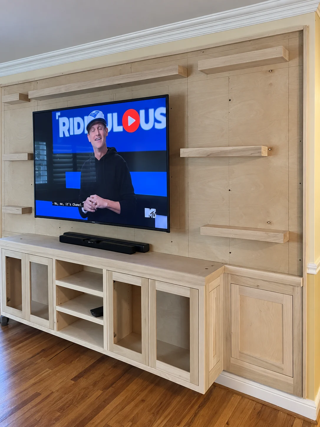 Media wall with glass-front cabinet doors and a paneled end; wood surfaces and surrounding wall remain unfinished